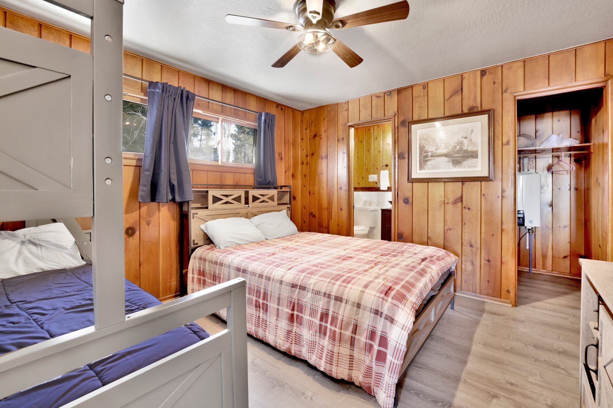 A cabin-style bedroom with a bunk bed, a single bed, wooden walls, a ceiling fan, a window with curtains, and a framed picture on the wall.