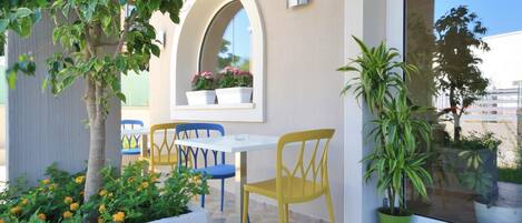 A patio area with a white table and yellow chairs, a potted plant, and a window with a view of the sky and buildings.