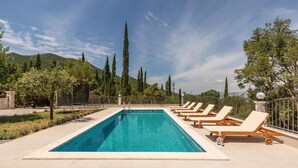 Ein rechteckiger Pool mit Liegestühlen am Rand, umgeben von Bäumen und einer Bergkulisse.