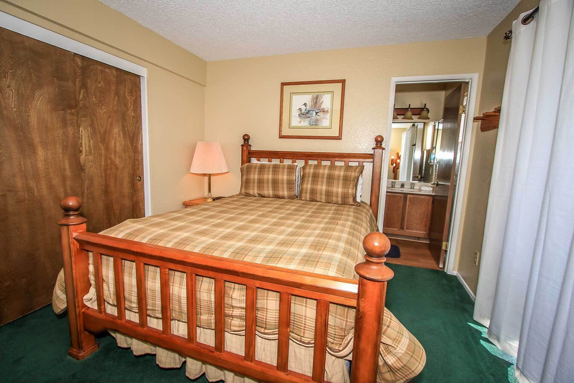Un dormitorio con cama de madera, una lámpara de noche, un cuadro enmarcado en la pared y un baño visible a través de una puerta abierta.
