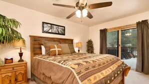 A bedroom with a large bed, wooden headboard, two bedside lamps, a nightstand, a plant, a framed picture, a ceiling fan, and a view of a snowy deck through a sliding glass door. - Swiss Delight #2244 (Big Bear Lake)