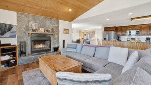 A cozy living room with a stone fireplace, a wooden ceiling, and a modern kitchen in the background. - I Love View #1475 (Big Bear City)