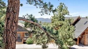A log cabin with a sloped roof, surrounded by pine trees, and a view of distant mountains. - I Love View #1475 (Big Bear City)