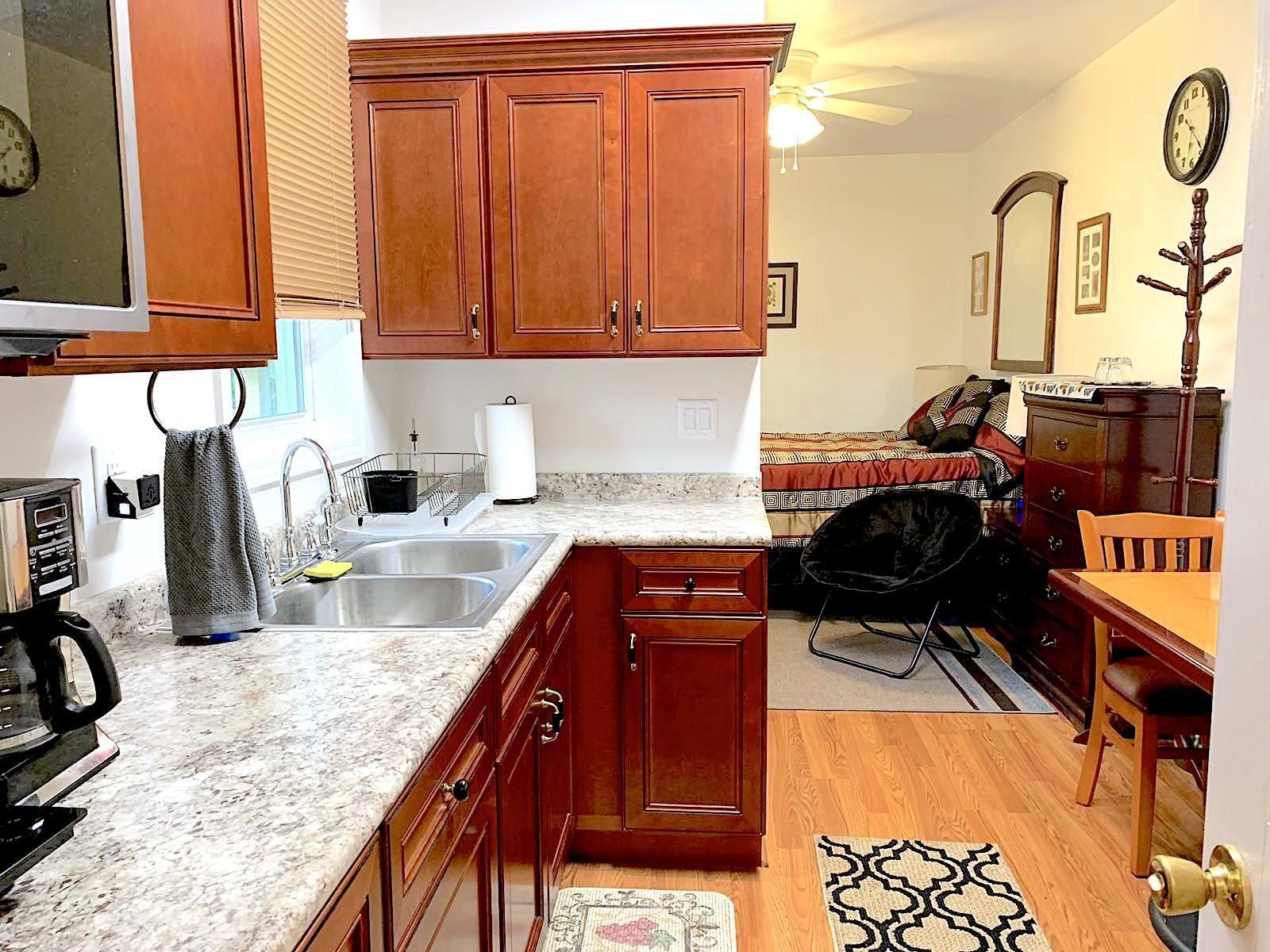 A compact kitchen with wooden cabinets, a sink, and a countertop. There is a dining area with a table and chairs, and a small rug on the floor.