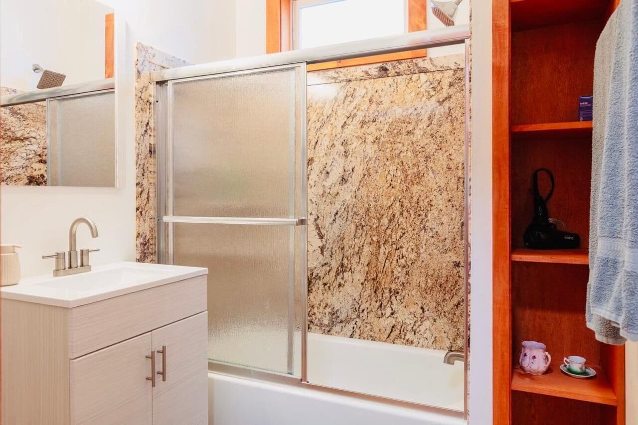 Un bany amb un lavabo blanc, un mirall, un espai de dutxa i una prestatgeria de fusta amb una tovallola.