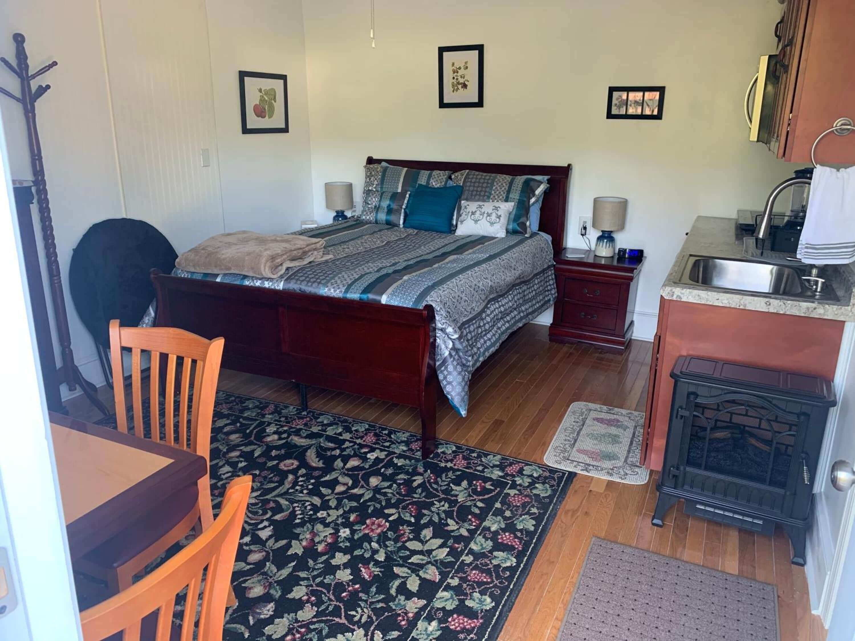 A bedroom with a wooden bed, a nightstand, a desk, a chair, a rug, a stove, and a sink.