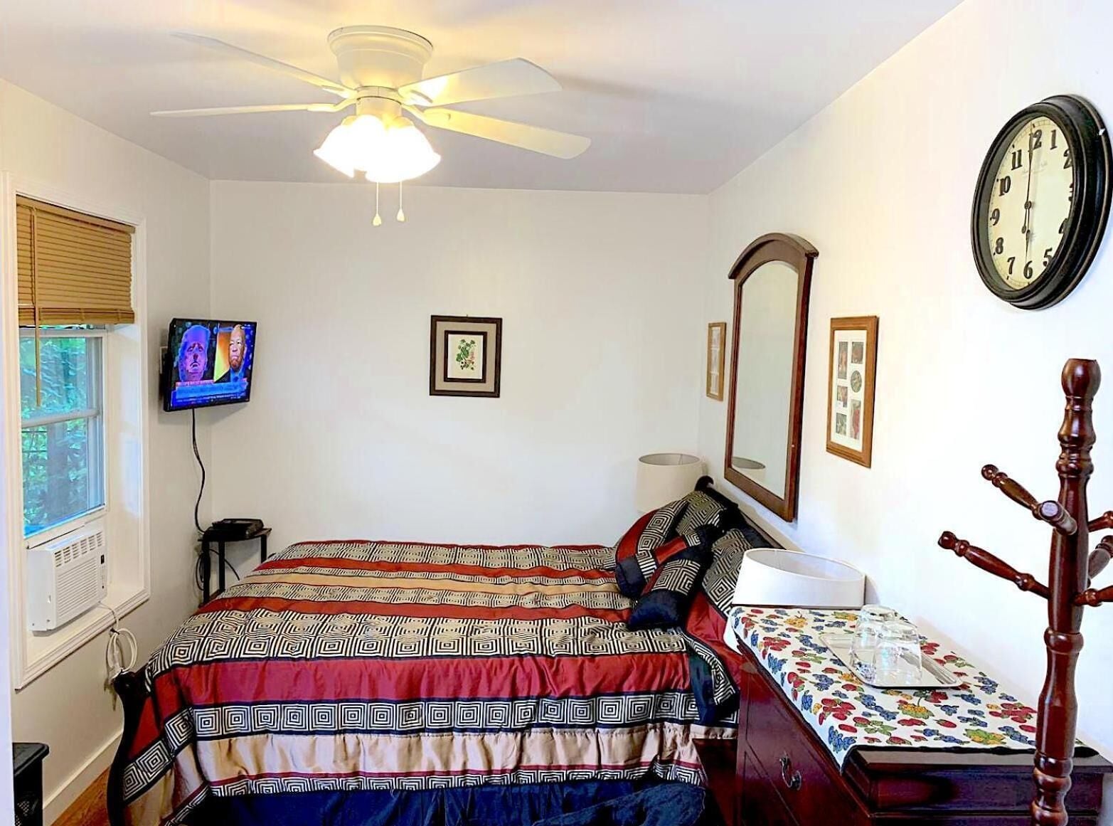 Un dormitorio con cama, ventilador de techo, televisor, ventana con persianas, mesita de noche con lámpara y un reloj de pared.