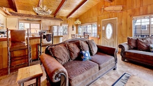 Ein gemütliches Blockhaus-Interieur mit Holzwänden, einer großen Holzbalkendecke, einer Couch mit gemusterten Kissen, einem Esstisch und einer Küchenzeile.