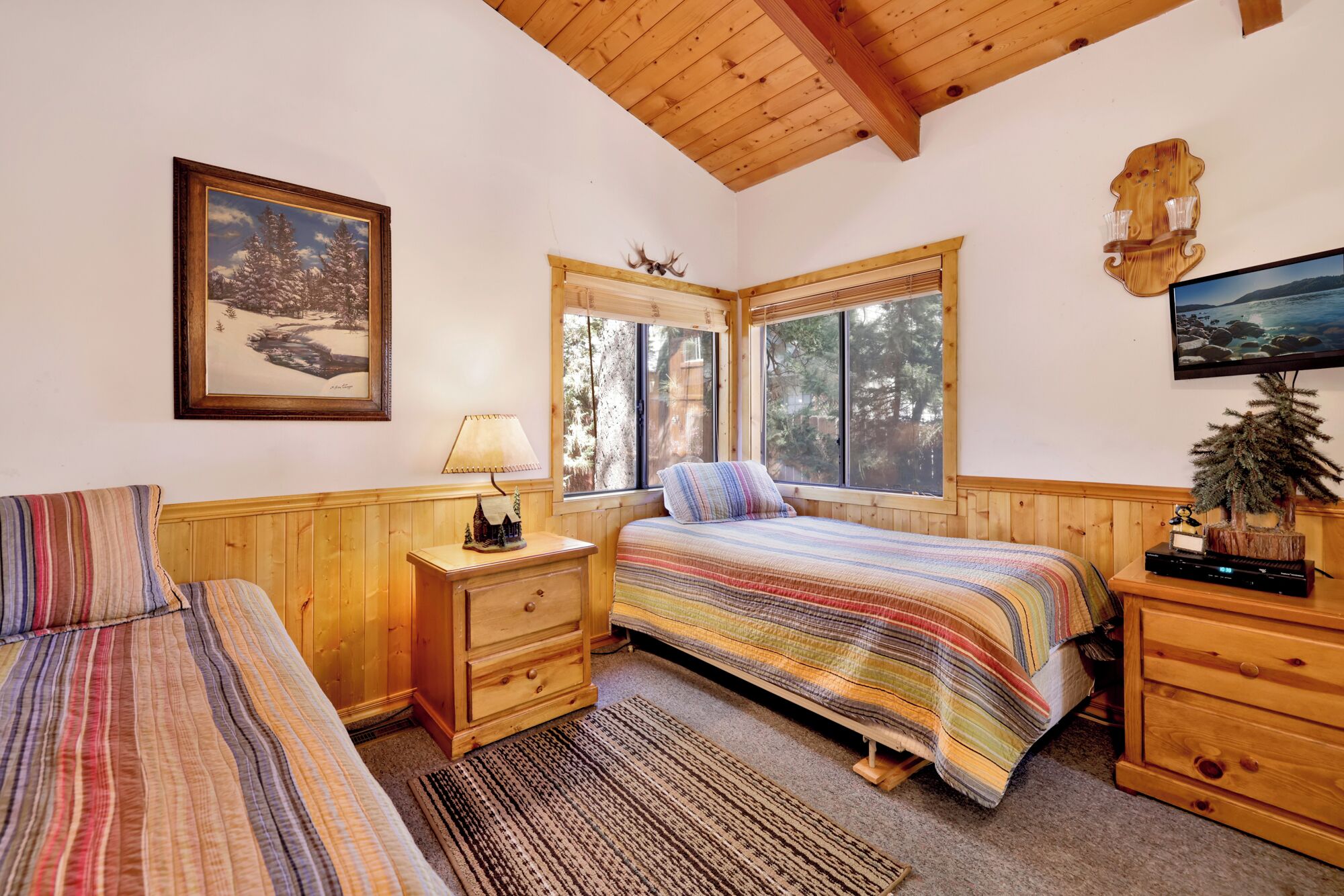 A cozy bedroom with two beds, wooden walls, a window with a view of trees, a lamp, and a small dresser.