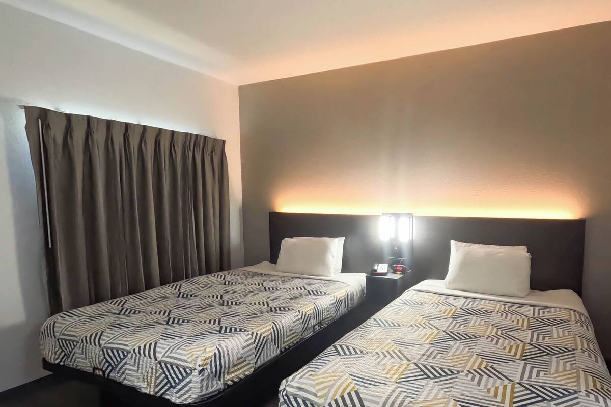 Une chambre d’hôtel avec deux lits, une table de chevet avec une lampe et une grande fenêtre avec des rideaux.