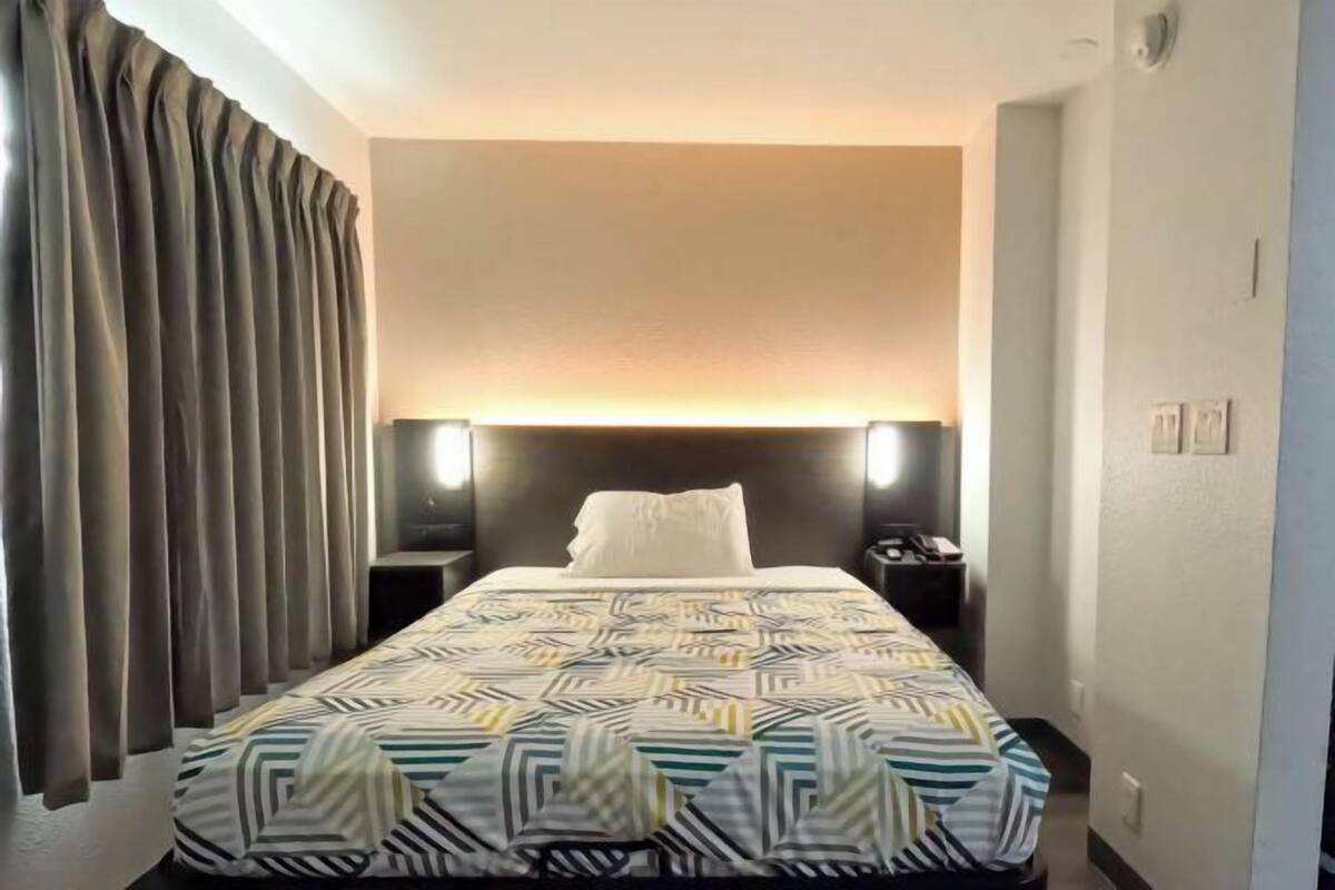 Une chambre d’hôtel avec un lit, des rideaux, une table de chevet et une lampe fixée au mur.
