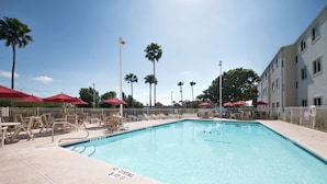 Una piscina rectangular con un cartel de prohibido bucear, rodeada de sillones de descanso y sombrillas rojas, con palmeras y un edificio al fondo.