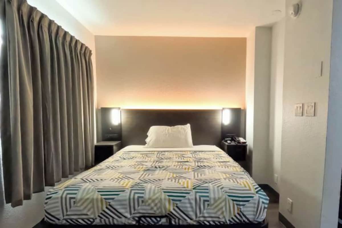 A hotel room with a bed, curtains, a nightstand, and a wall-mounted light.