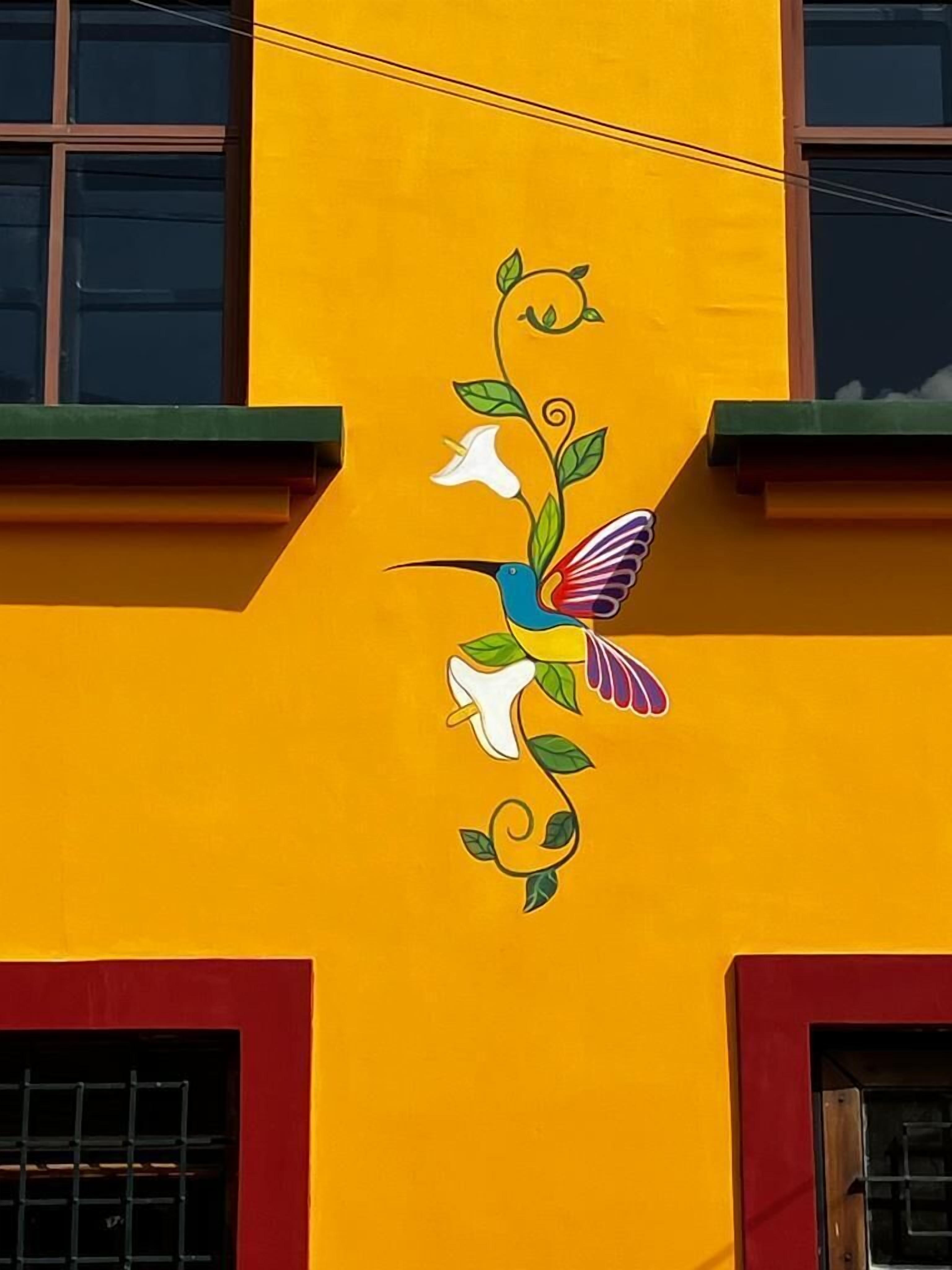 A building with a vibrant yellow facade and a colorful mural of a hummingbird and flowers.