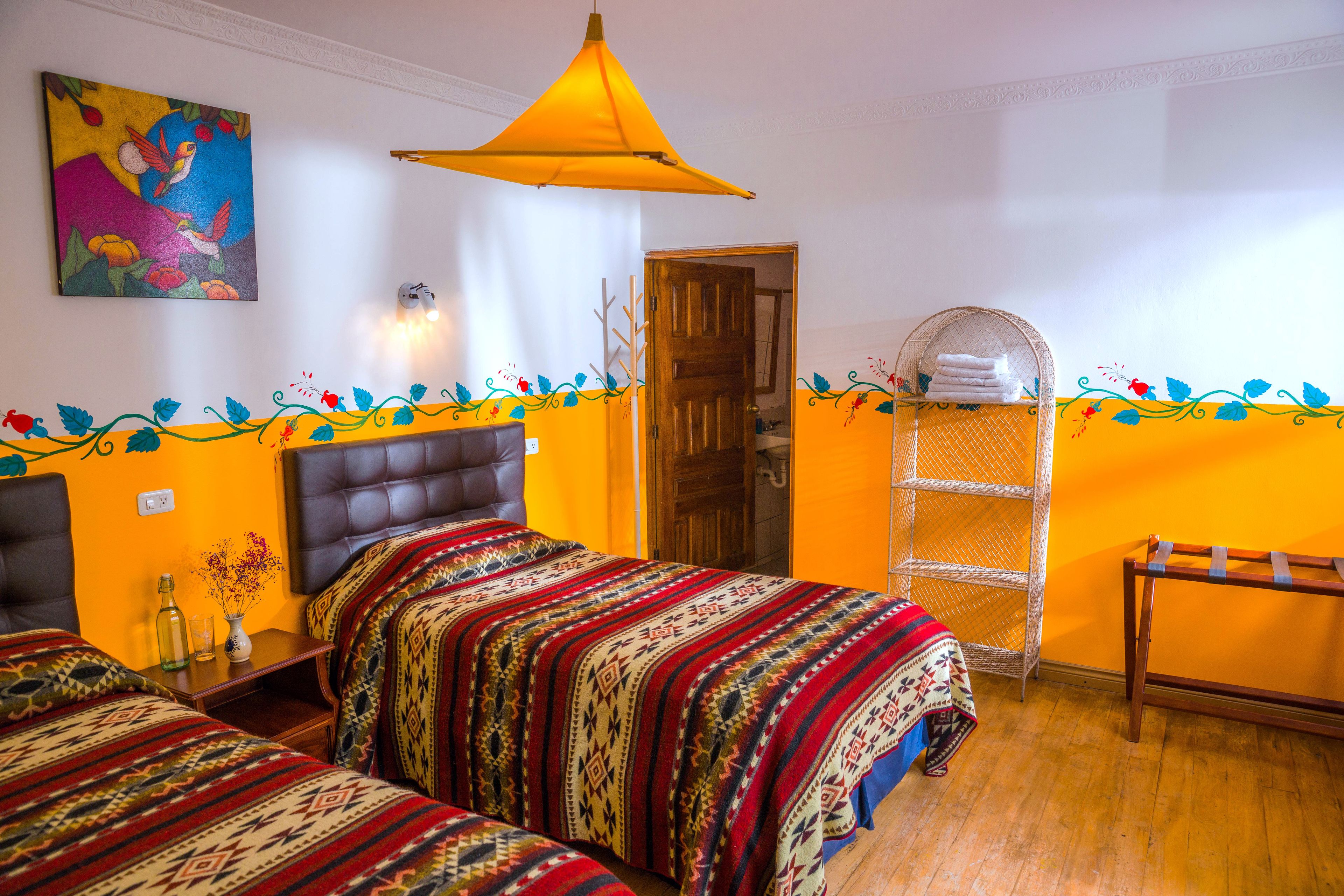 Ein Zimmer mit zwei Betten, einem Hängestuhl, einem Regal mit Handtüchern und einer farbenfrohen Wanddekoration.