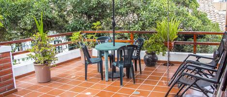 Un patio con una mesa redonda y sillas, rodeado de plantas en macetas y un paraguas grande.