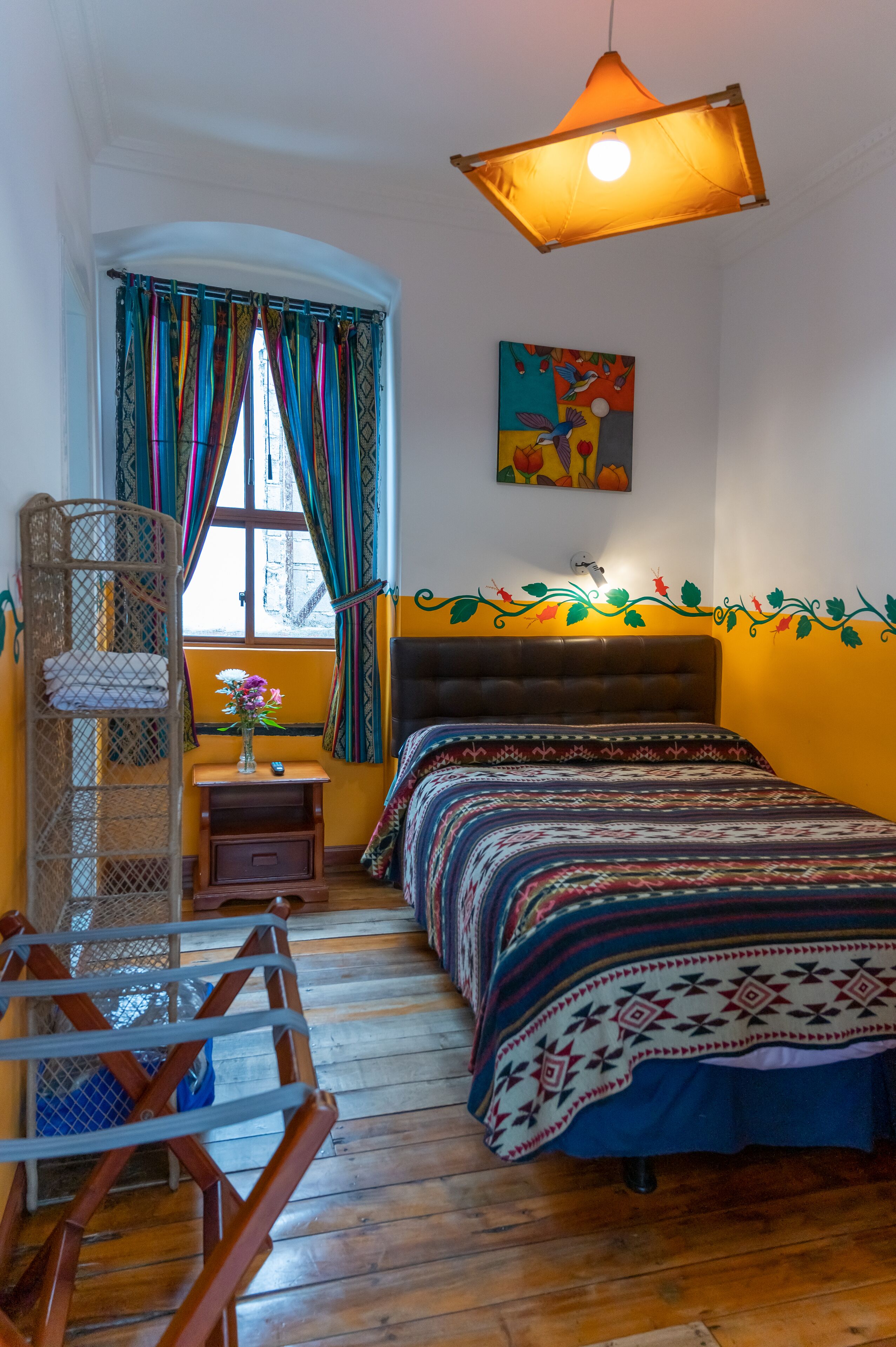 A room with a bed, a window with colorful curtains, a hanging light fixture, and a wall decoration.