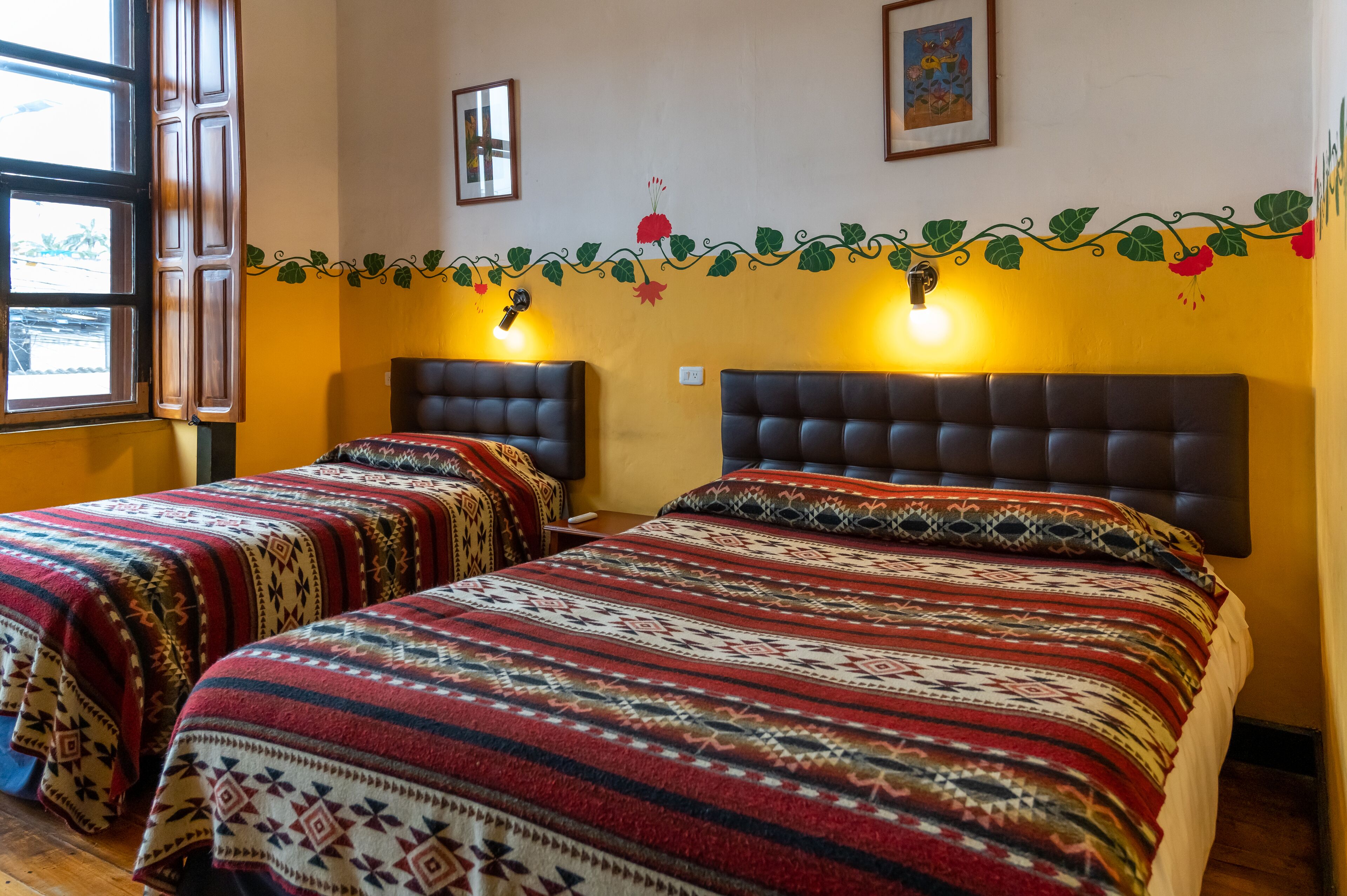 Une chambre avec deux lits, chacun agrémenté de draps à motifs, une fenêtre avec des rideaux et des décorations murales comprenant un motif de vigne et des tableaux encadrés.