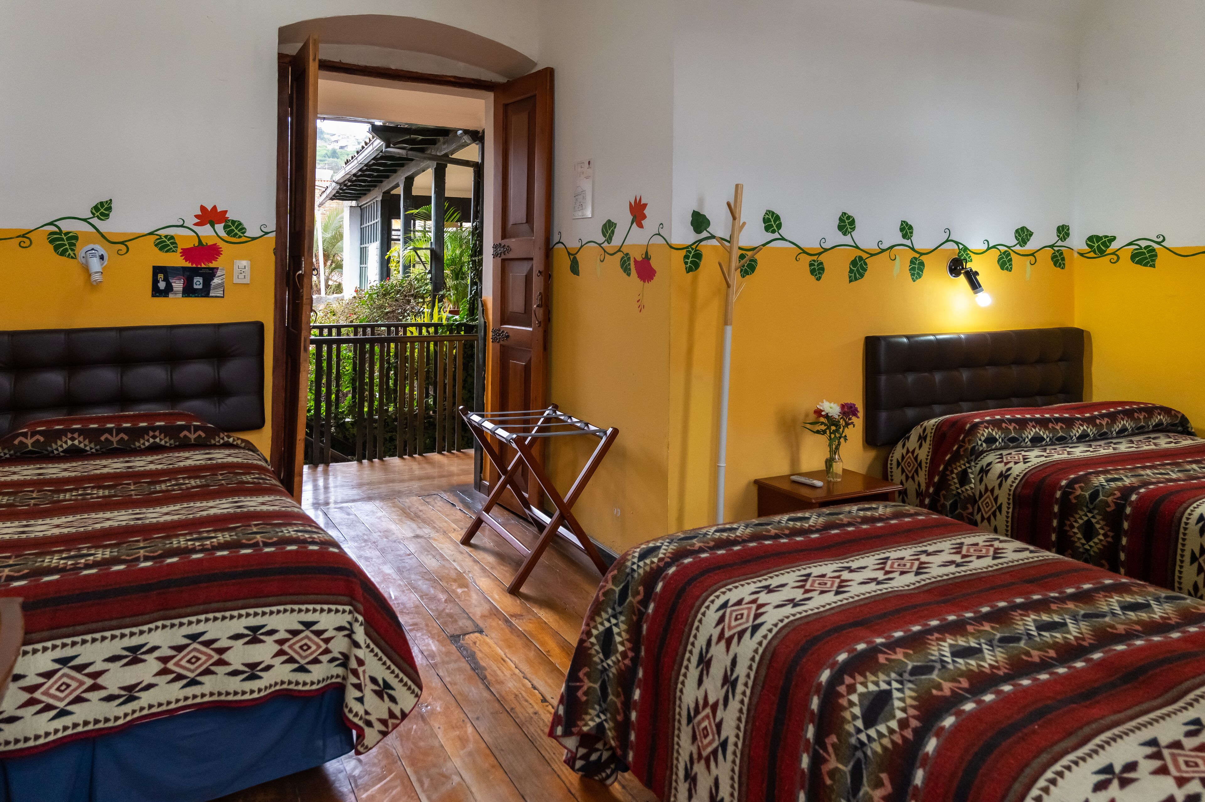 Una habitación con dos camas, piso de madera, balcón y una pared decorada con enredaderas y flores.