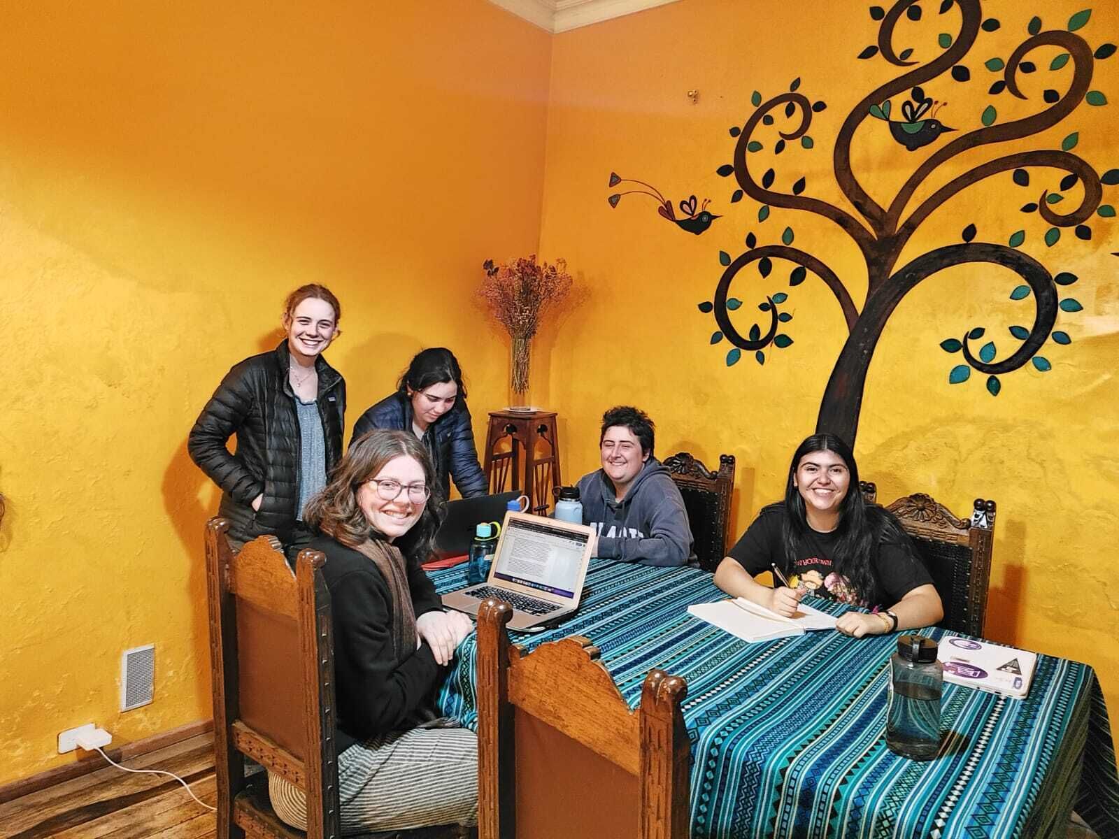 Un grupo de personas sentadas alrededor de una mesa de madera en una habitación con paredes amarillas y un mural de un árbol.