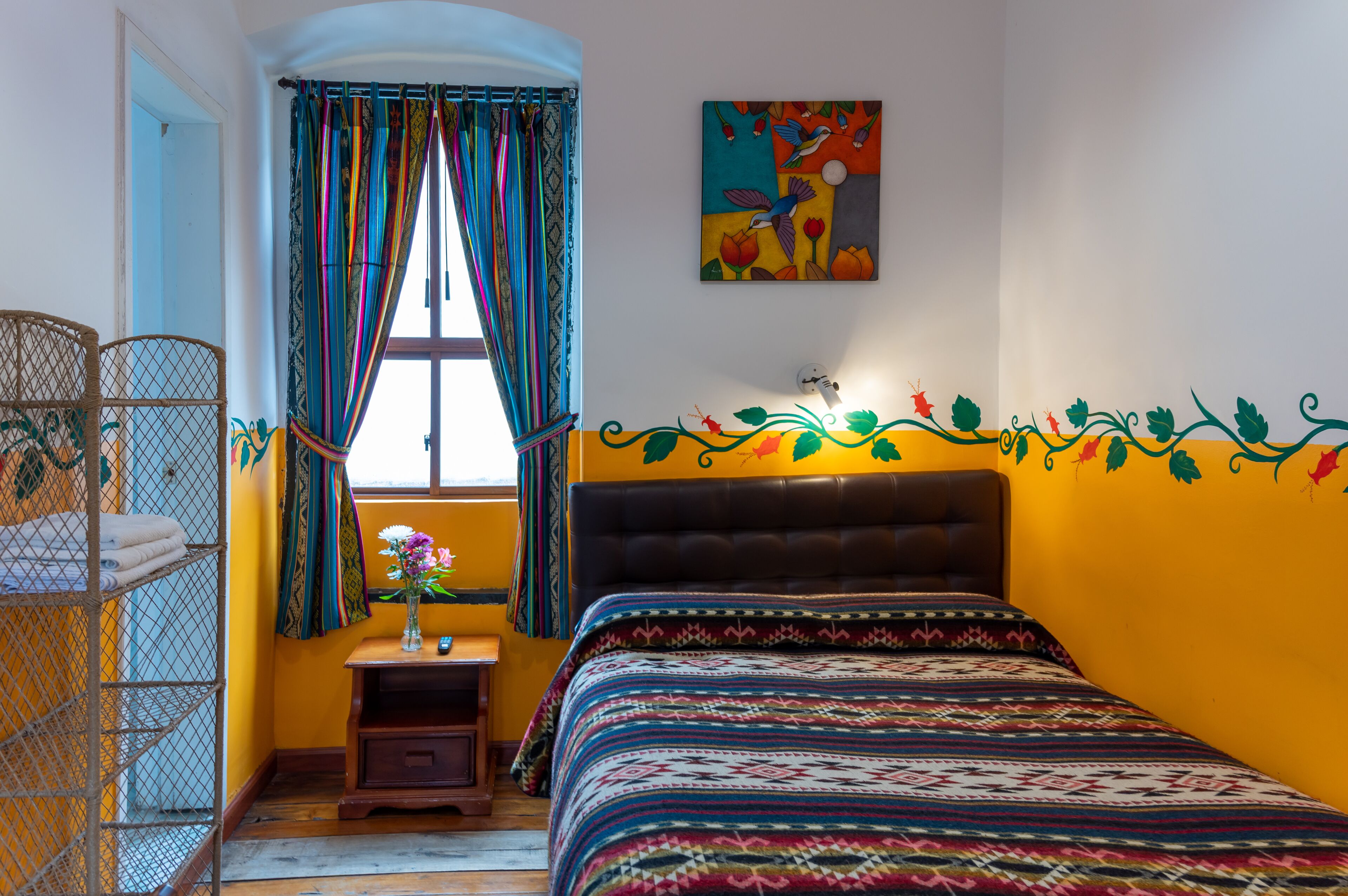 A room with a bed, a window with colorful curtains, a wall with a floral mural, and a small table with a vase.