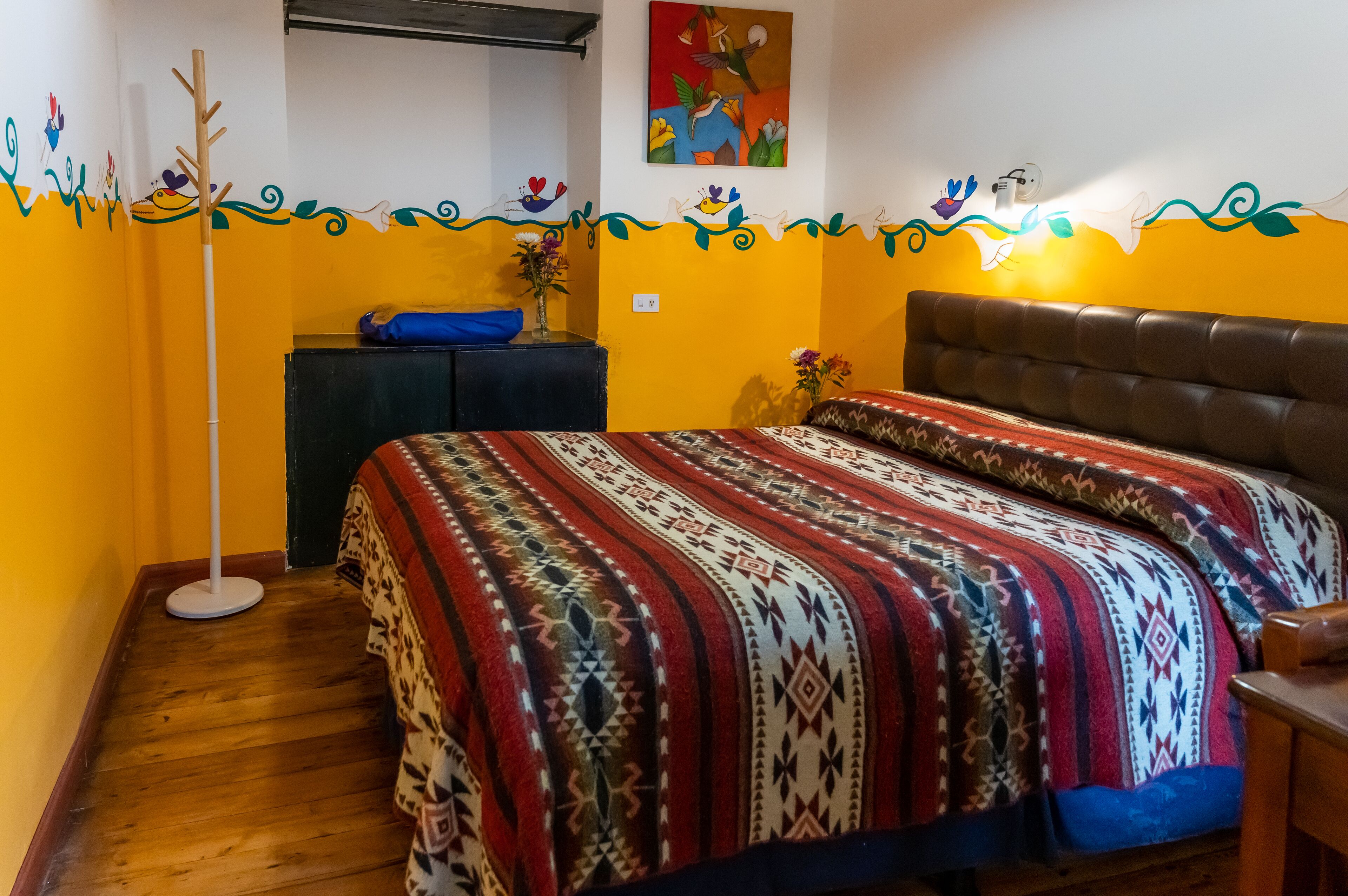 A room with a patterned bedspread, a wooden floor, a desk with a chair, and a wall with colorful artwork.