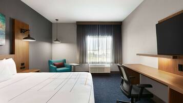 A hotel room with a bed, a desk, a chair, a TV, a window with curtains, and an air conditioning unit.
