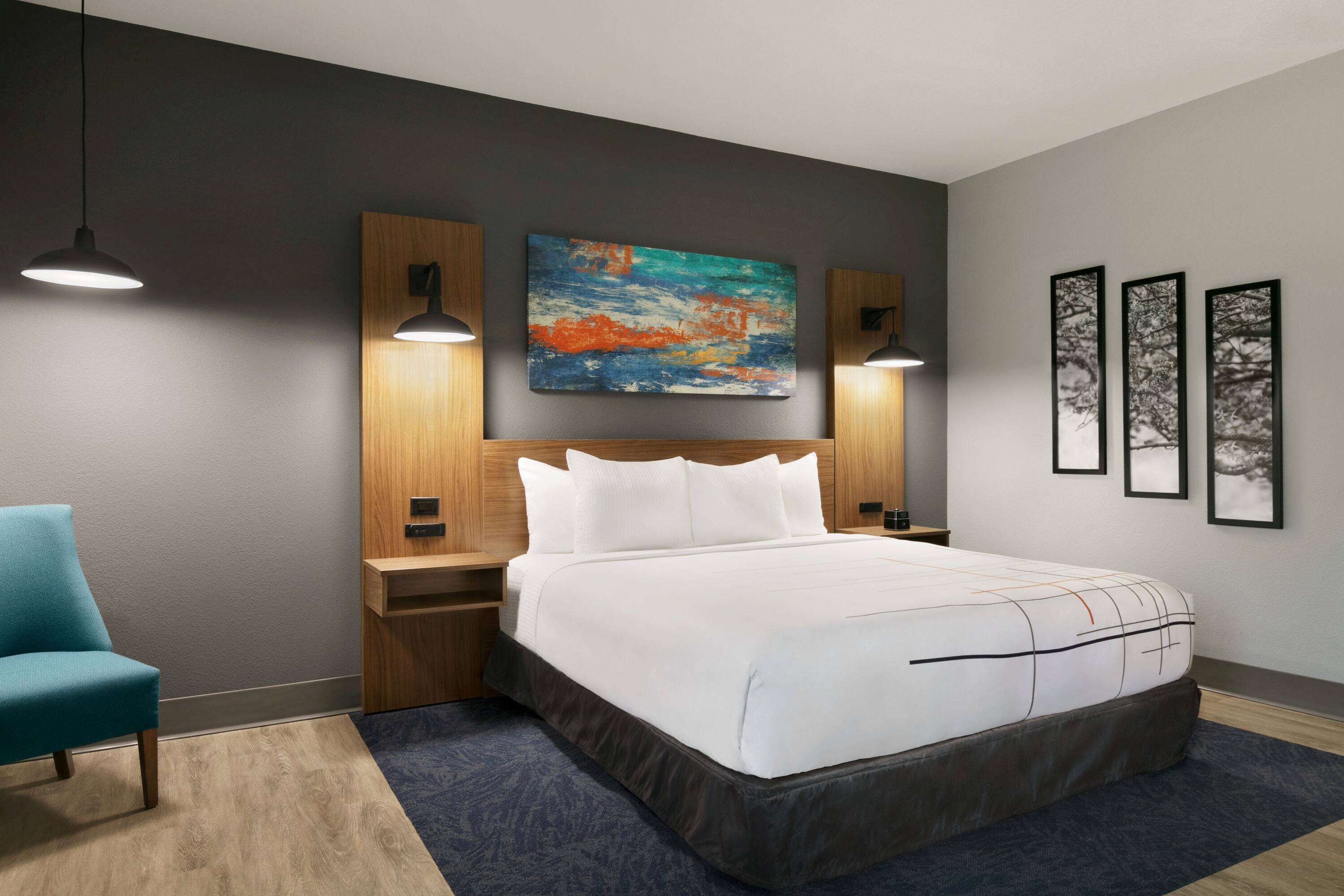 A modern hotel room with a large bed, a blue armchair, a wooden headboard, and a colorful abstract painting above the bed.
