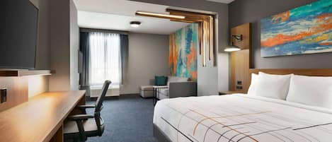 A hotel room with a large bed, a desk, a chair, a small table, and a painting on the wall.