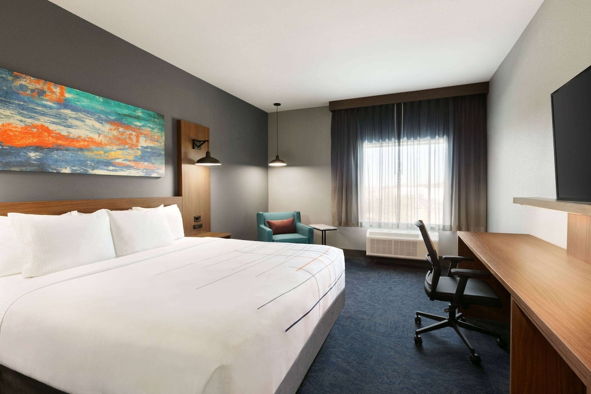 Habitación de hotel con una cama grande, un escritorio, una silla, una mesita, una lámpara, un cuadro y una ventana con cortinas.