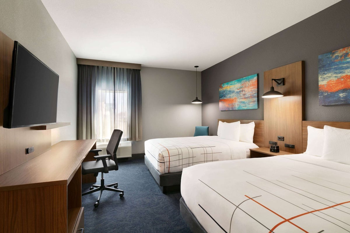 Habitación de hotel con escritorio, dos camas, televisor de pantalla plana, obras de arte en las paredes y ventana con cortinas.