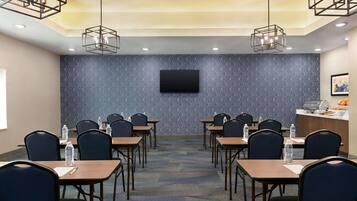 A conference room with a patterned blue wall, a flat-screen TV, and a buffet area with fruits and snacks.