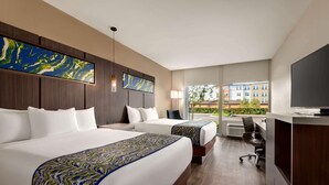 Habitación de hotel con dos camas, un escritorio y un amplio ventanal con vistas a edificios y zonas verdes.