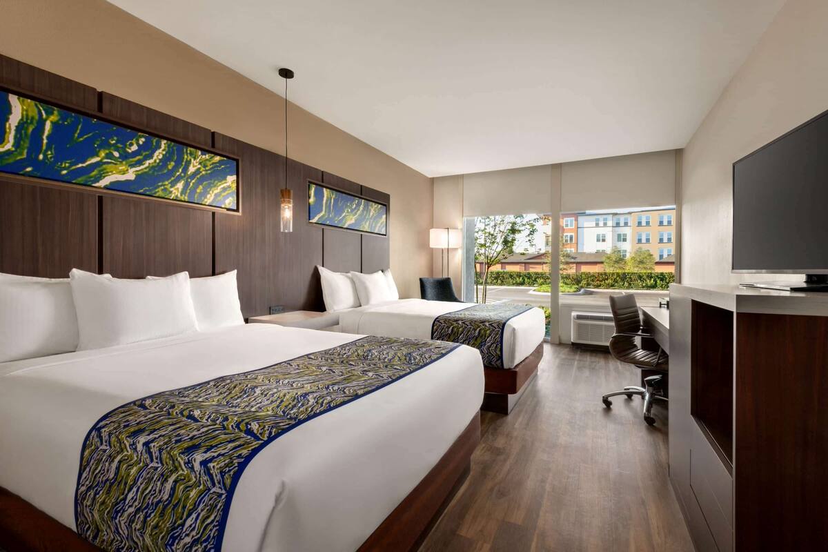 Habitación de hotel con dos camas, un escritorio y un amplio ventanal con vistas a edificios y zonas verdes.
