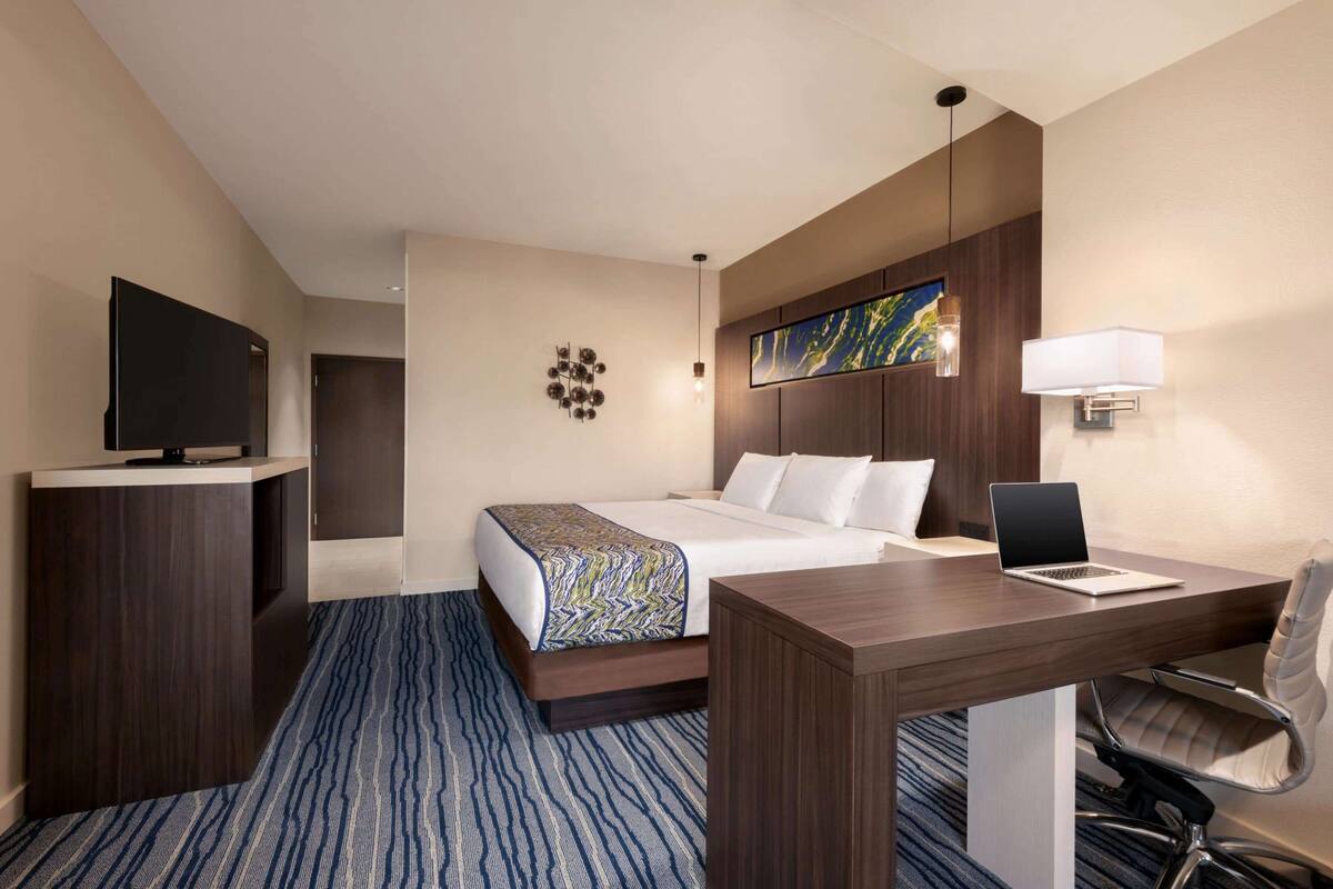 Una habitación de hotel con una cama, un escritorio con una computadora portátil, un televisor y una obra de arte en la pared.