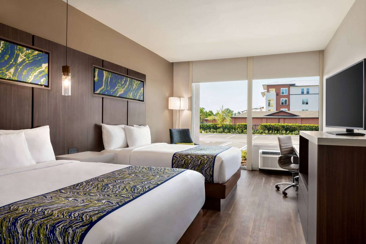 Habitación de hotel con dos camas, un escritorio, una silla, un televisor y un ventanal con vistas a edificios.