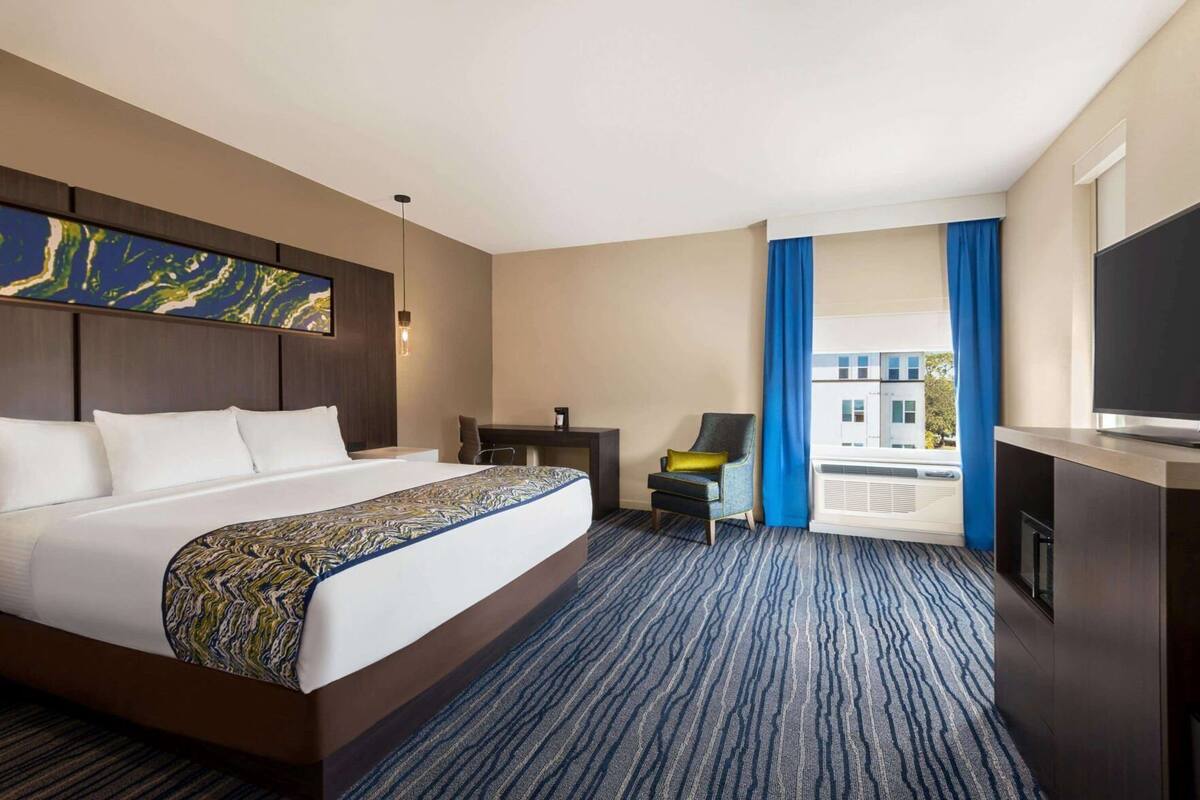 Una habitación de hotel con una cama grande, un escritorio, una silla, un televisor y un ventanal con cortinas.