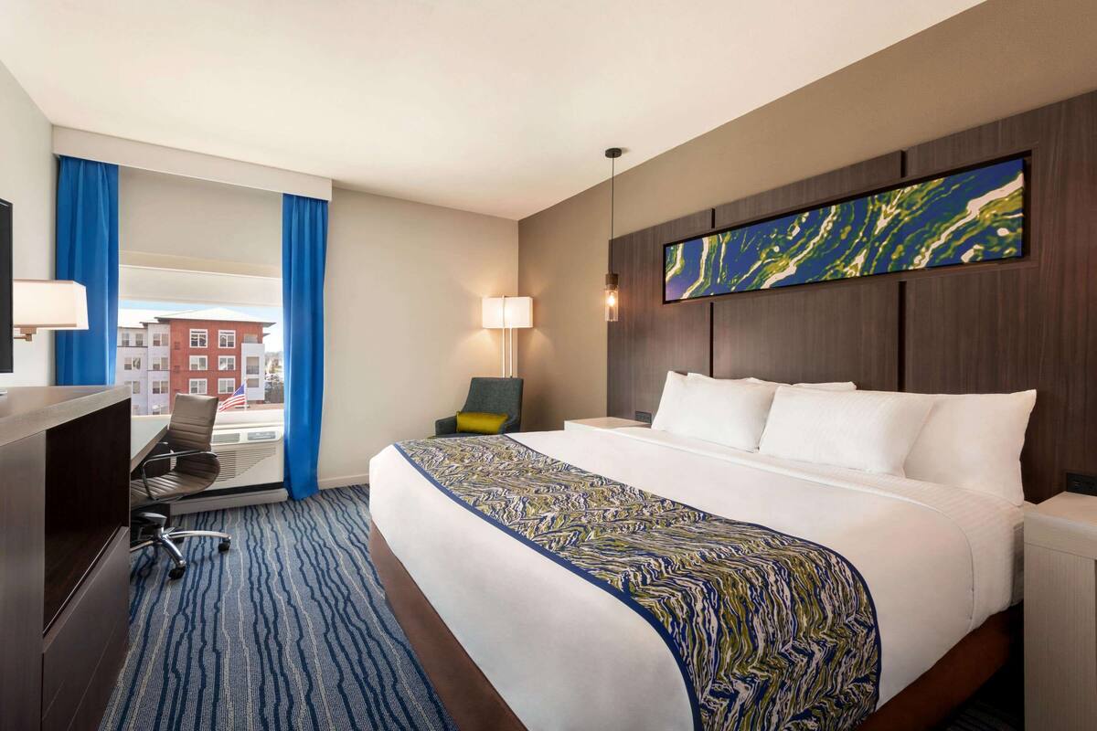 Una habitación de hotel con una cama grande, un escritorio y una silla. Hay una ventana con cortinas azules y vistas a edificios.