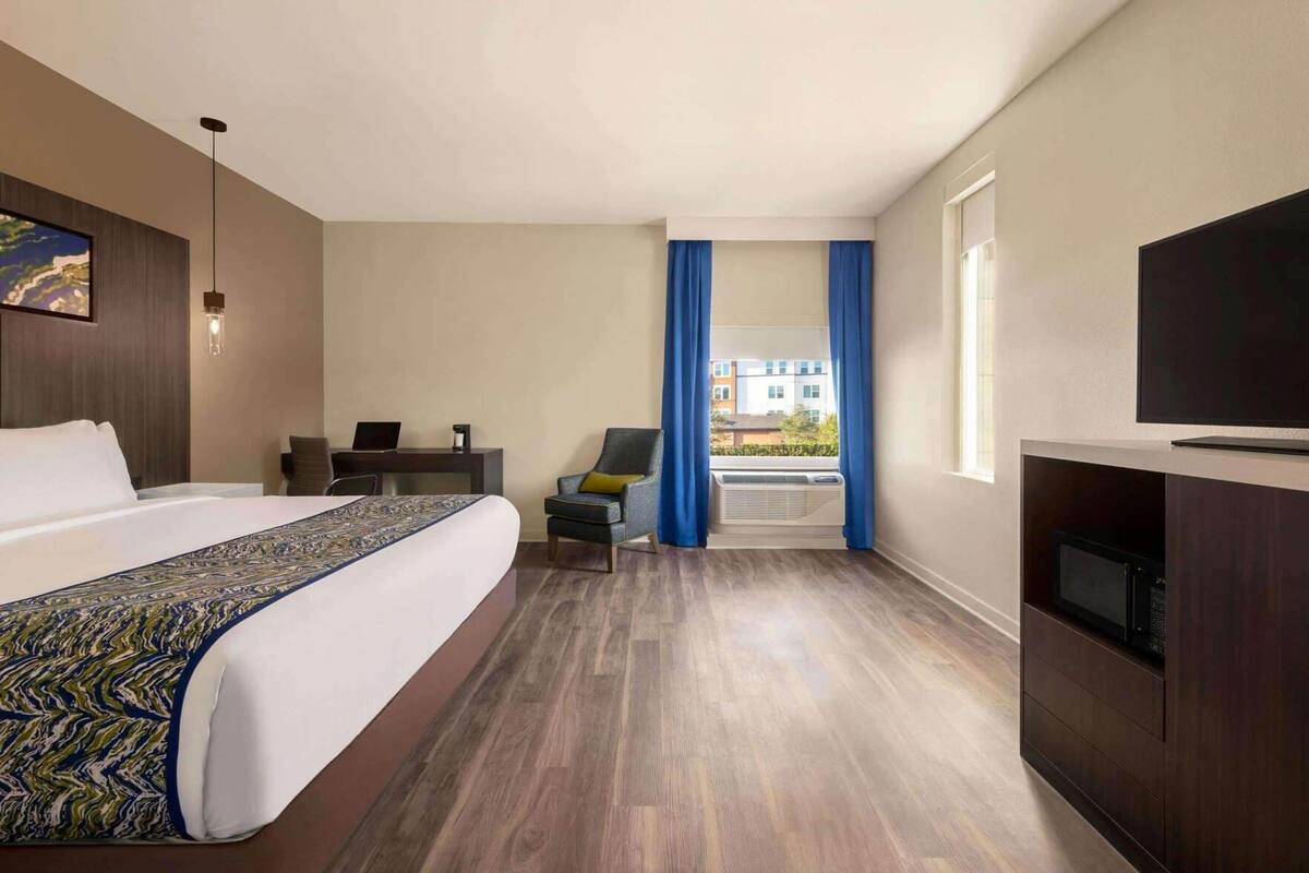 Habitación de hotel con una cama grande, un escritorio con una computadora portátil, una silla, un televisor y vistas a edificios a través de una ventana.