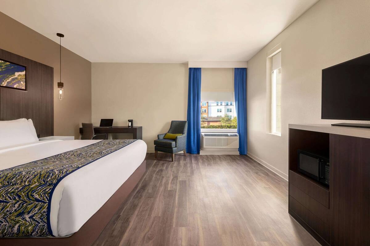 Habitación de hotel con una cama grande, un escritorio, una silla, un televisor y vistas a edificios a través de la ventana.