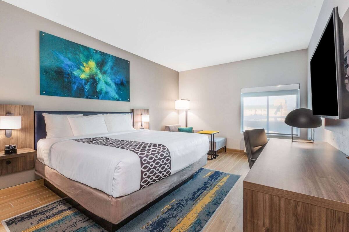 Une chambre d’hôtel avec un grand lit, des tables de chevet, un bureau, une chaise, une télévision et un grand tableau abstrait accroché au mur.