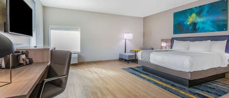 Una habitación de hotel con cama, escritorio, silla, televisor, lámpara y un cuadro en la pared.