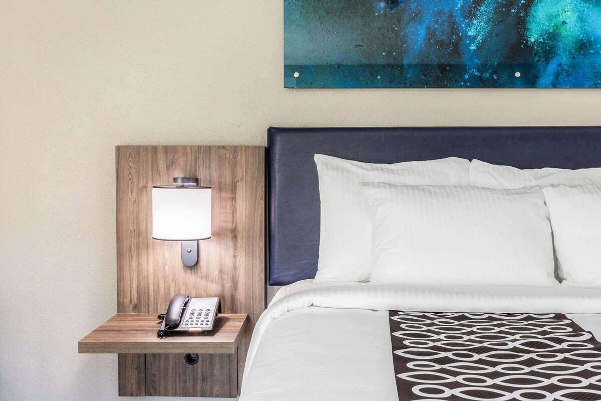 Une chambre d’hôtel avec une table de chevet en bois, une lampe, un téléphone, un lit avec une literie blanche et un grand tableau abstrait accroché au mur.