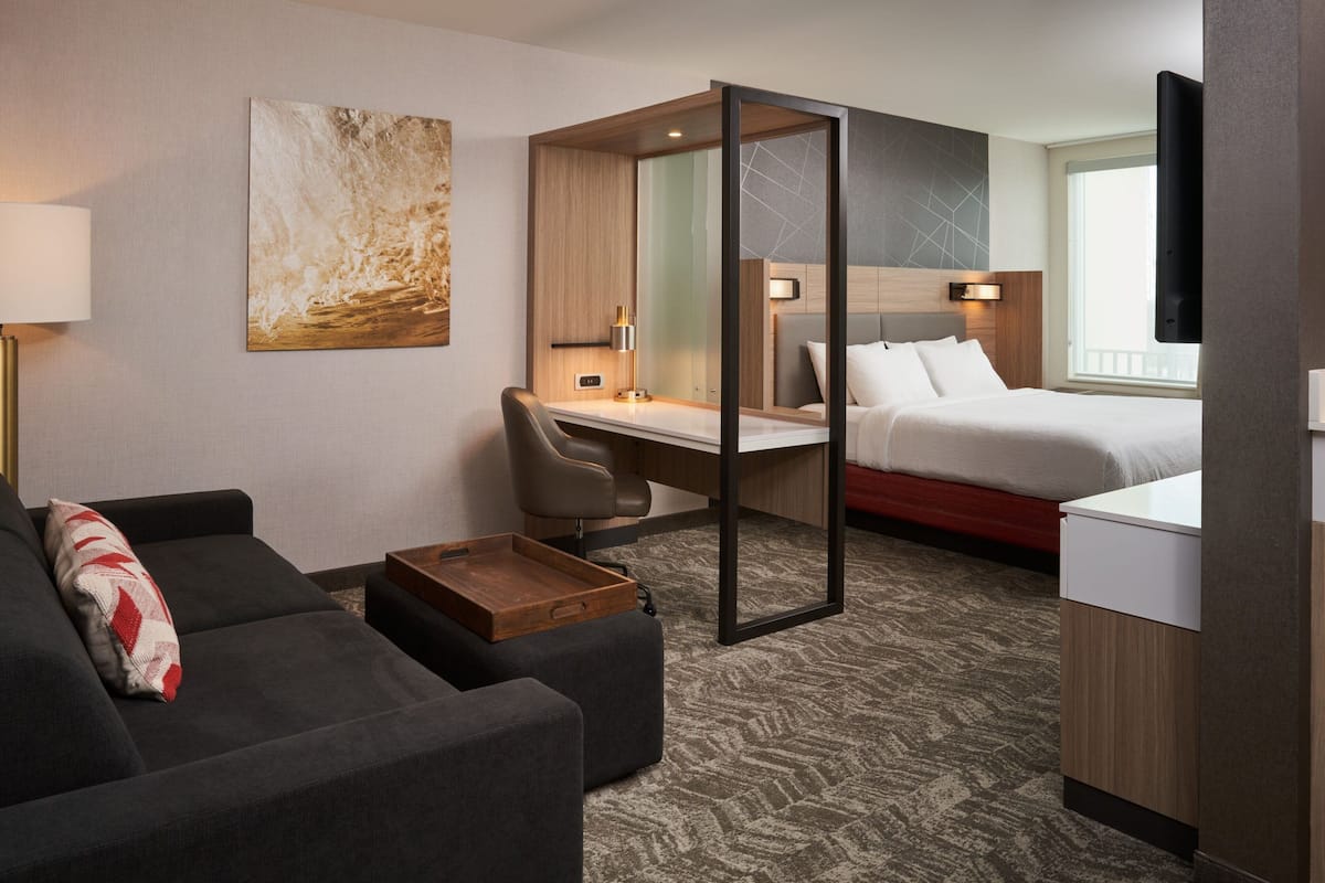A hotel room with a bed, a desk, a chair, a sofa, a TV, and a painting on the wall.
