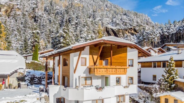 A modern chalet with a wooden balcony, surrounded by snow-covered trees and a mountainous backdrop.