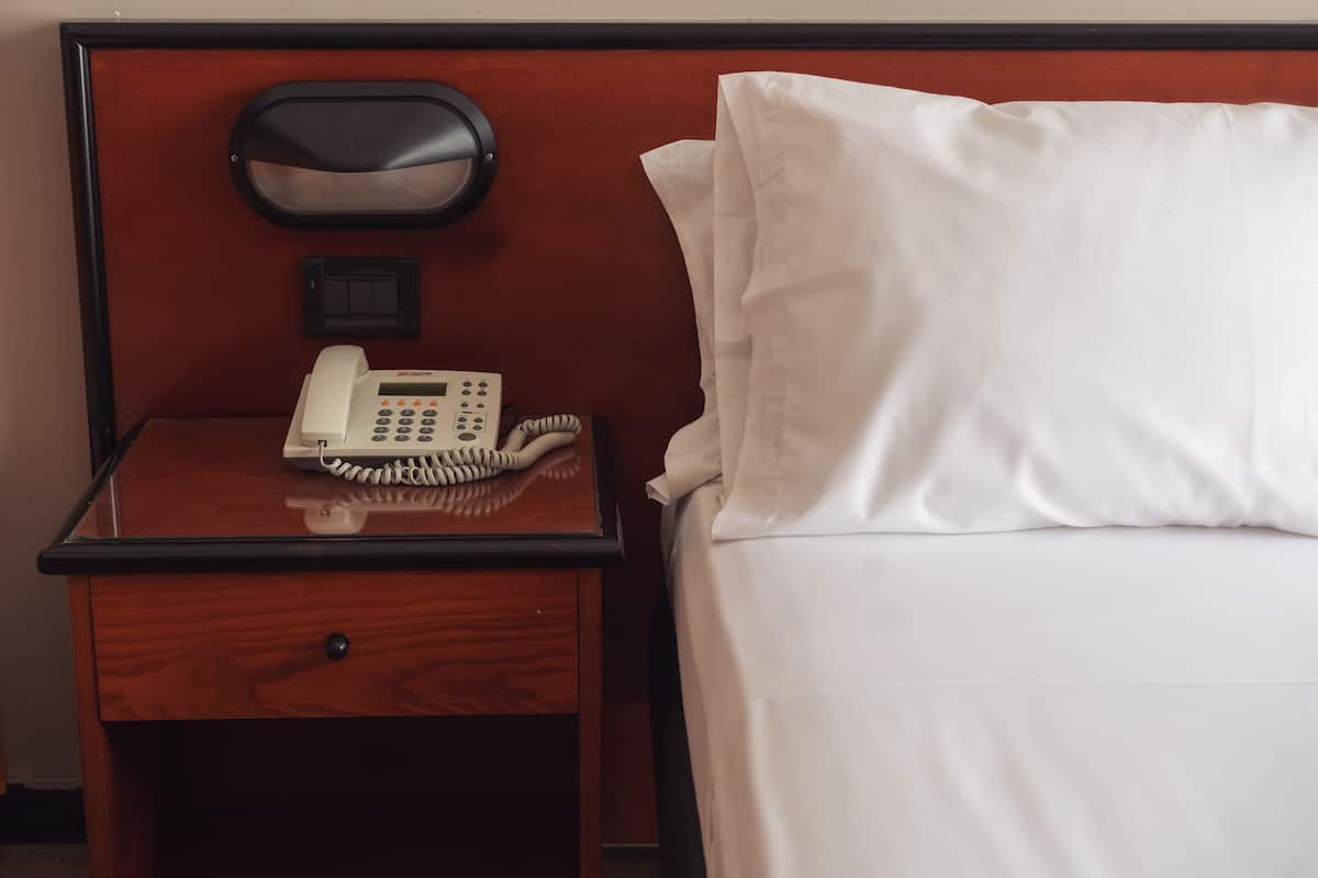 A hotel room with a wooden nightstand, a telephone, and a white bed with a pillow.