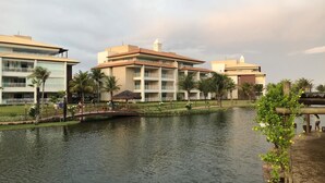 A hotel with multiple floors, a water body in front, and palm trees. - Golf Ville Resorts Suites (Aquiraz)