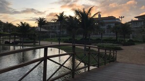A wooden bridge over a calm water body, surrounded by palm trees and buildings with a sunset sky in the background. - Golf Ville Resorts Suites (Aquiraz)