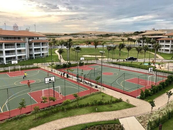 Una cancha polideportiva con canchas de baloncesto y tenis, rodeada de zonas ajardinadas con palmeras y vistas a edificios lejanos.