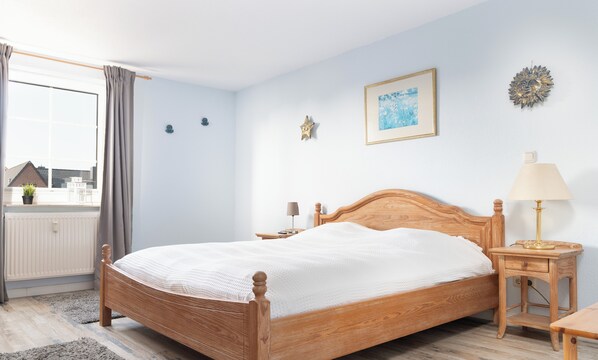 A bedroom with a wooden bed, a bedside table with a lamp, a window with curtains, and a wall with a framed picture and a star decoration. - Vacation apartment Parkpension W16 (Laboe)